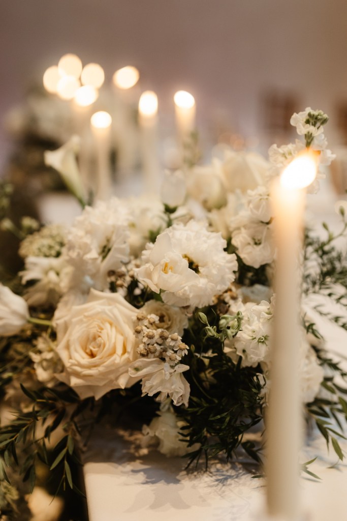 A romantic, close-up shot of a wedding table centrepiece with white and cream flowers, surrounded by the warm, soft glow of tall, elegant candles.