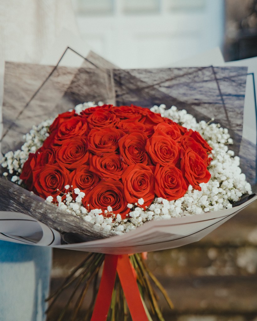 A close-up of a bouquet of vibrant red roses, elegantly arranged and surrounded by delicate white baby's breath flowers. The bouquet is wrapped in a combination of white and black paper with a subtle geometric pattern. The soft, natural lighting highlights the rich colour of the roses. Perfect for weddings, special events, or as a thoughtful gift.