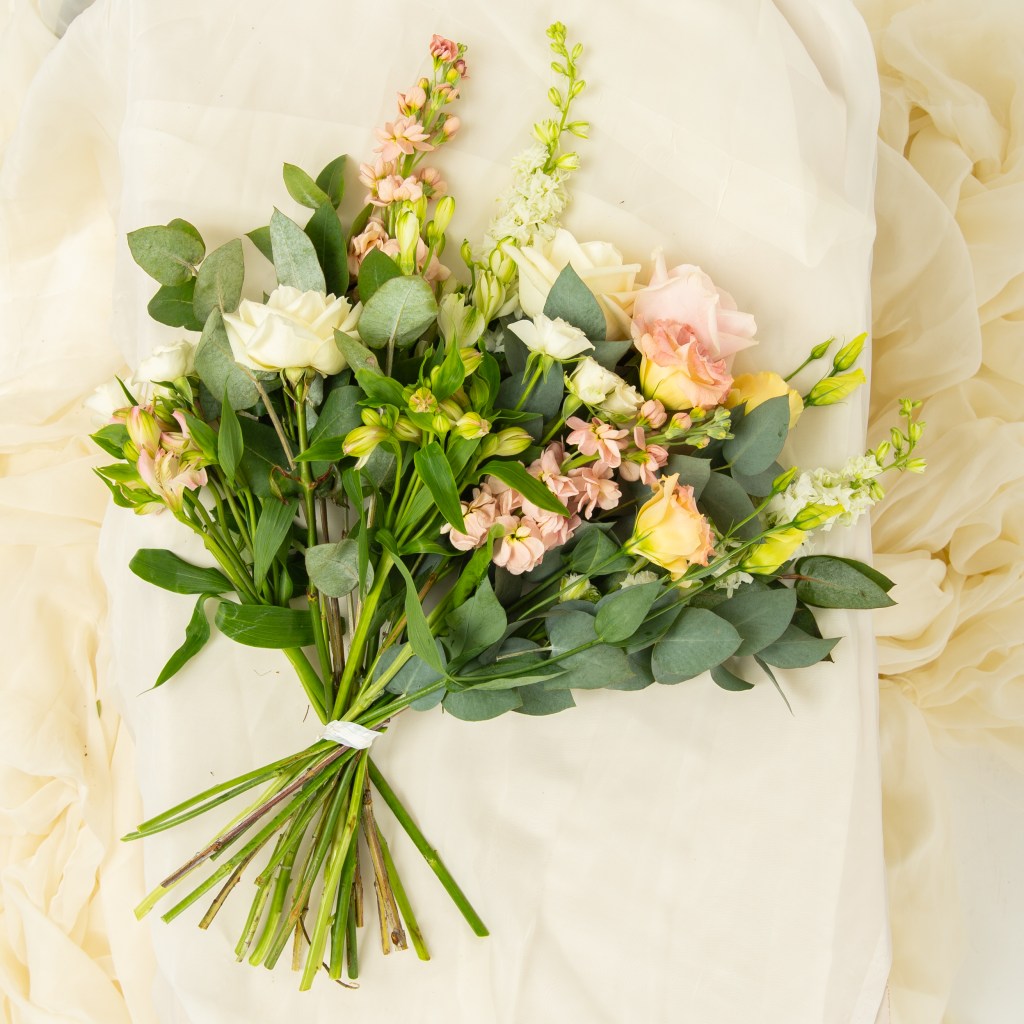 A bouquet of soft peach, white, and pale yellow roses is arranged with delicate greenery and eucalyptus leaves. The stems are tied together and laid on light cream fabric, creating an elegant and minimalist display.